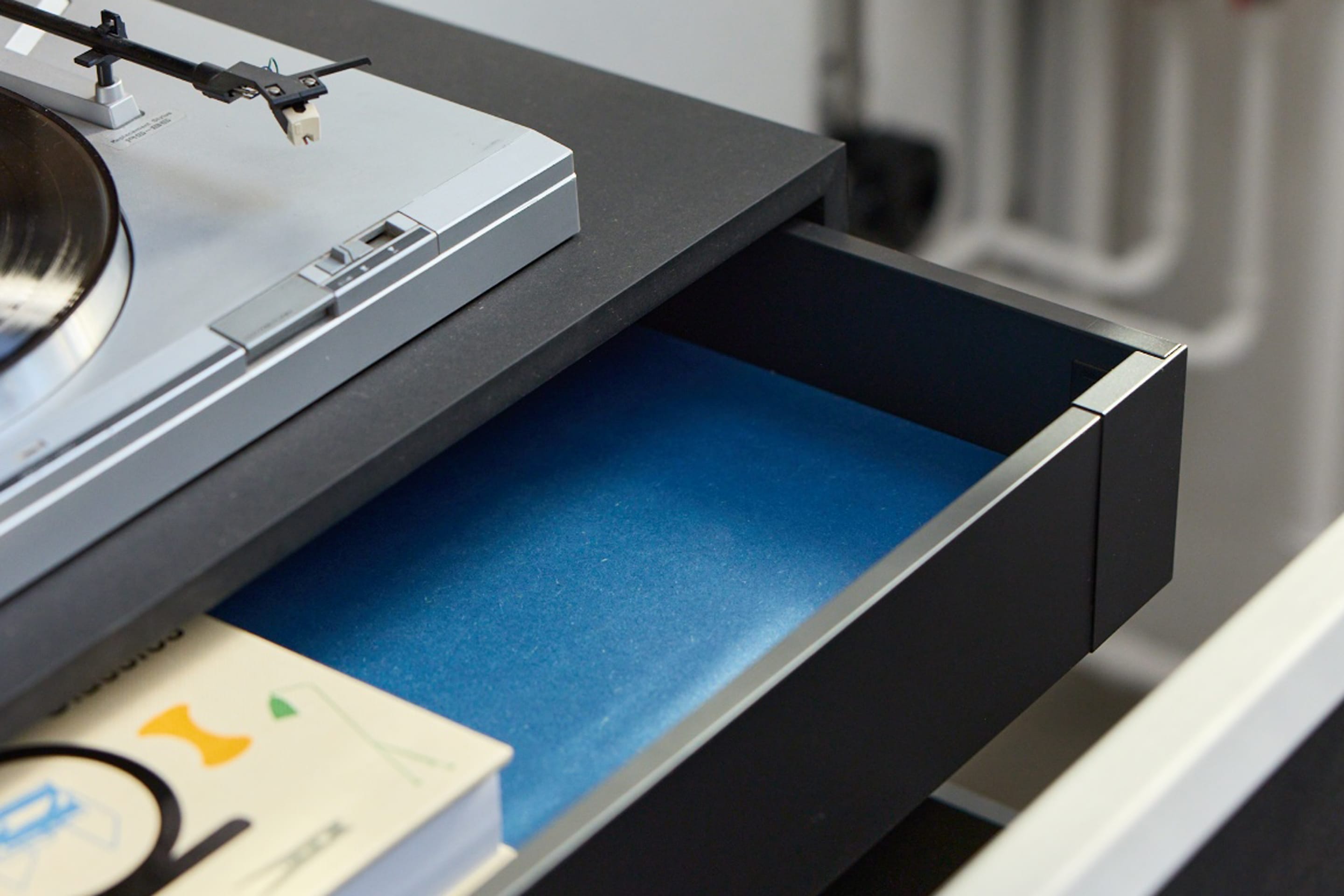 The Corpus in Schwarz mit geöffneter Schublade mit blauem Boden, darauf ein Plattenspieler. Holzfaserwerkstoff, durchgefärbt.